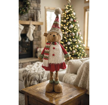 Weihnachts-Stoffbär in festlicher Kleidung auf einem Holztisch, im Hintergrund ein geschmückter Weihnachtsbaum und ein Kamin.
