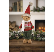 Weihnachts-Teddybär stehend mit rot-grünem Strickpullover, roter Mütze und weißem Schal vor einem Kamin mit Tannenzweigen.