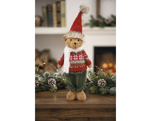 Weihnachts-Teddybär stehend mit rot-grünem Strickpullover, roter Mütze und weißem Schal vor einem Kamin mit Tannenzweigen.