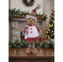 Dekorativer Plüschbär in winterlicher Kleidung mit Strickmütze und Schal vor weihnachtlicher Kulisse mit Kamin und Tannenbaum.