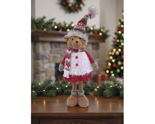 Dekorativer Plüschbär in winterlicher Kleidung mit Strickmütze und Schal vor weihnachtlicher Kulisse mit Kamin und Tannenbaum.