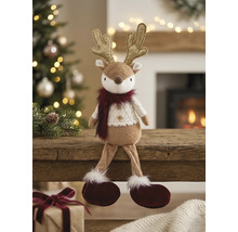 Plüsch-Rentier mit goldenem Geweih und weißem Strickpullover auf einem Holzbalken in einem hellen Raum mit Weihnachtsbaum.