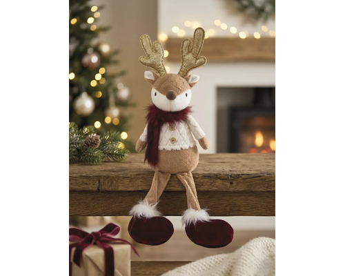 Plüsch-Rentier mit goldenem Geweih und weißem Strickpullover auf einem Holzbalken in einem hellen Raum mit Weihnachtsbaum.