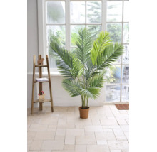 Künstliche Palme in einem Topf in einem hellen Raum mit Fliesenboden, großem Fenster und einem dekorativen Holzregal.