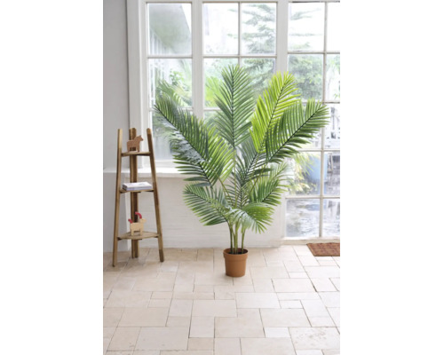 Künstliche Palme in einem Topf in einem hellen Raum mit Fliesenboden, großem Fenster und einem dekorativen Holzregal.