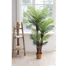 Künstliche Palme mit mehreren Stämmen im Kunststofftopf in einem hellen Raum neben einem Holzregal vor einem Fenster.