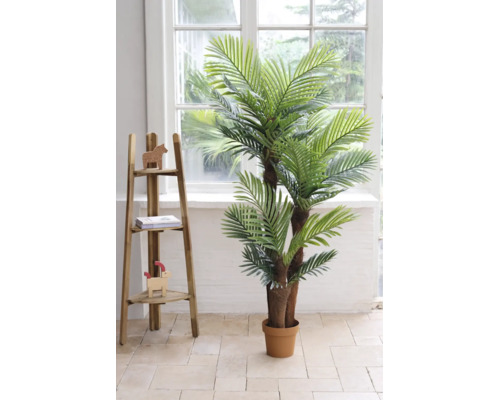 Künstliche Palme mit mehreren Stämmen im Kunststofftopf in einem hellen Raum neben einem Holzregal vor einem Fenster.