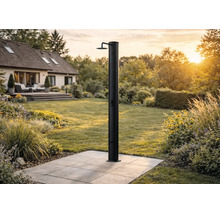 Schwarze Solardusche auf einer gepflasterten Terrasse in einem weitläufigen, hellen Garten mit Wohnhaus im Hintergrund.