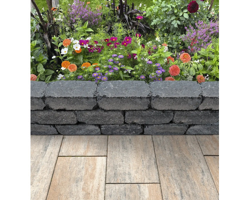 Niedrige Gartenmauer aus dunkelgrauen strukturierten Mauersteinen vor einem bunten Blumenbeet und hellen Terrassenplatten.