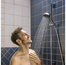 Mann unter einer verchromten Handbrause an einer Duschstange in einem Badezimmer mit blauen und weißen Fliesen.