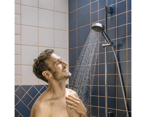 Mann unter einer verchromten Handbrause an einer Duschstange in einem Badezimmer mit blauen und weißen Fliesen.