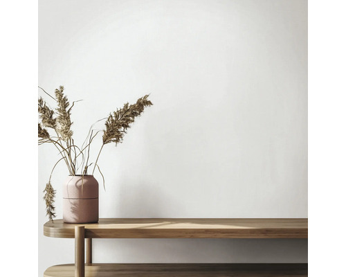 Minimalistischer Innenraum mit einer Holzbank und einer strukturierten Vase mit getrocknetem Schilfgras vor einer hellen Wand.