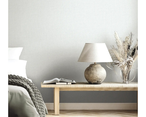 Hellgraue Vliestapete in Leinenoptik in einem hellen Schlafzimmer mit Holzbank, Tischlampe, Trockenblumen und Bett mit Strickdecke.