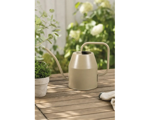 Moderne Metallgießkanne auf einem Holztisch im Garten mit kleiner Pflanze und Gartenhandschuhen in einer hellen Atmosphäre.