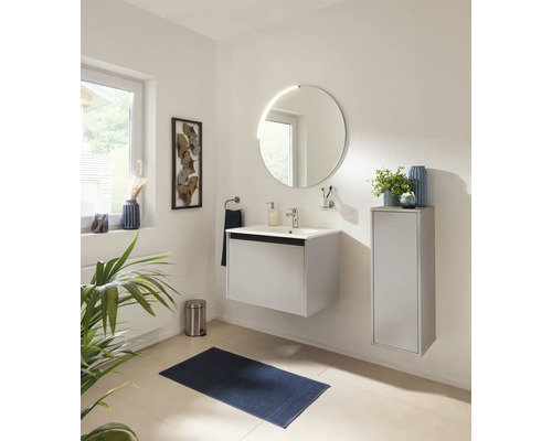 Modernes, helles Badezimmer mit weißem Waschtisch, rundem Spiegel und Seitenschrank. Mit Badematte und Treteimer aus Edelstahl.