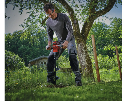 Mann bedient einen Erdbohrer mit Akkubetrieb im Garten zum Bohren von Löchern für Zaunpfosten unter einem Baum.