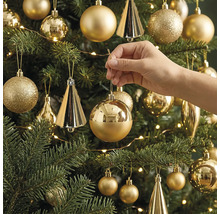 Eine Hand hängt eine glänzende, runde Weihnachtskugel an einen geschmückten Christbaum mit verschiedenen Ornamenten und einer Lichterkette.