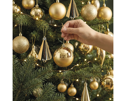 Eine Hand hängt eine glänzende, runde Weihnachtskugel an einen geschmückten Christbaum mit verschiedenen Ornamenten und einer Lichterkette.