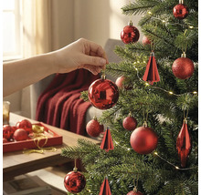Hand hängt eine rote Kugel an einen geschmückten Weihnachtsbaum mit Lichtern und verschiedenen roten Ornamenten in einem hellen Raum.