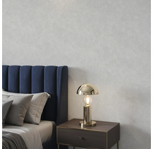 Goldfarbene Tischleuchte auf einem Nachttisch in einem Schlafzimmer mit blauem Polsterbett und grauen Kissen vor einer strukturierten Wand.
