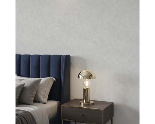 Goldfarbene Tischleuchte auf einem Nachttisch in einem Schlafzimmer mit blauem Polsterbett und grauen Kissen vor einer strukturierten Wand.