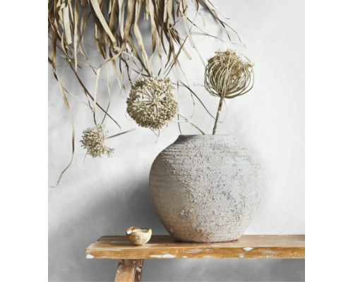Runde Keramikvase mit rauer Textur und getrockneten Blüten auf einer Holzbank vor einer hellen Wand.