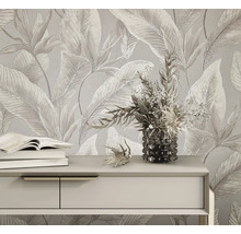 Helle Vliestapete mit großflächigem tropischem Blättermuster und Blüten hinter einem Sideboard mit Büchern und einer Blumenvase.
