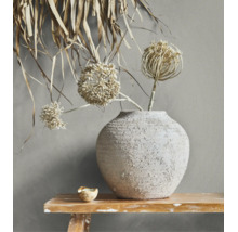 Rustikale Keramikvase mit strukturierter Oberfläche und getrockneten Blumen auf einer Holzbank vor einer Wand.