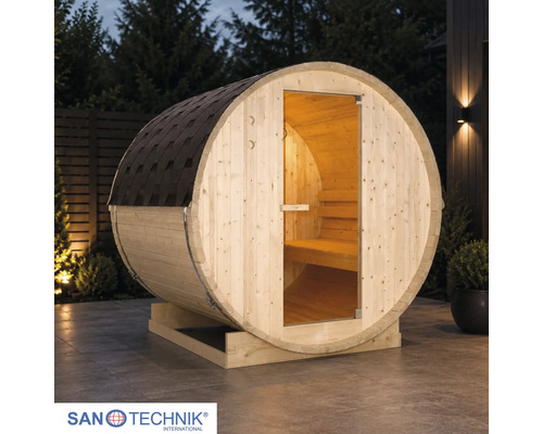 Sanotechnik Fasssauna aus Holz auf einer Terrasse bei Dämmerung mit Glastür, Innenbeleuchtung und Dachschindeln. Sanotechnik Logo.