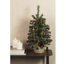 Dekorierter künstlicher Weihnachtsbaum mit LED-Beleuchtung und Jutesack-Sockel neben weißen Kerzen auf einem Tisch.