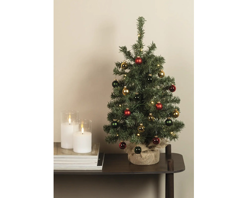 Dekorierter künstlicher Weihnachtsbaum mit LED-Beleuchtung und Jutesack-Sockel neben weißen Kerzen auf einem Tisch.