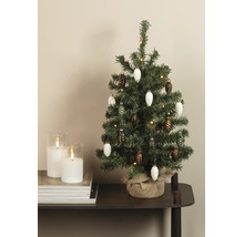 Kleiner künstlicher Weihnachtsbaum mit Zapfenanhängern, Lichterkette und Jutesack neben zwei weißen Kerzen im Glas auf einem Stapel Zeitschriften.