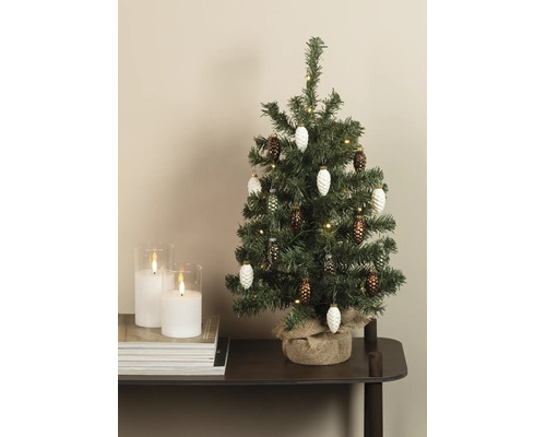 Kleiner künstlicher Weihnachtsbaum mit Zapfenanhängern, Lichterkette und Jutesack neben zwei weißen Kerzen im Glas auf einem Stapel Zeitschriften.