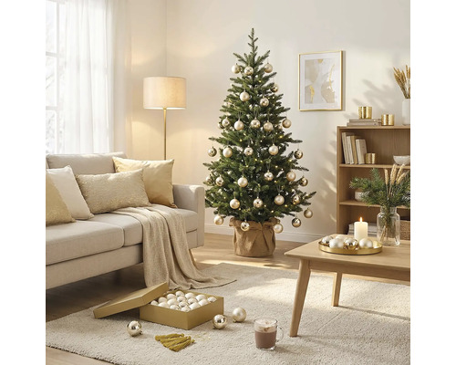 Geschmückter Weihnachtsbaum mit Kugeln und Lichterkette in einem hellen Wohnzimmer mit Sofa, Couchtisch und Teppich.