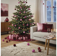 Festlich geschmückter Weihnachtsbaum in einem hellen Wohnzimmer mit rosa und burgunderroten Kugeln, Lichterkette, Geschenken und Sofa.