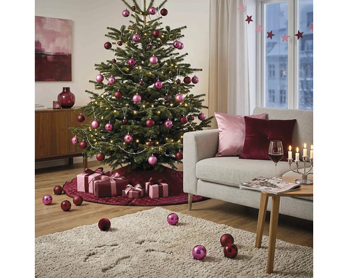 Festlich geschmückter Weihnachtsbaum in einem hellen Wohnzimmer mit rosa und burgunderroten Kugeln, Lichterkette, Geschenken und Sofa.