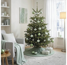 Geschmückter Weihnachtsbaum mit Kugeln und Lichtern im hellen Wohnzimmer neben einem Sessel mit Strickdecke und Geschenken.