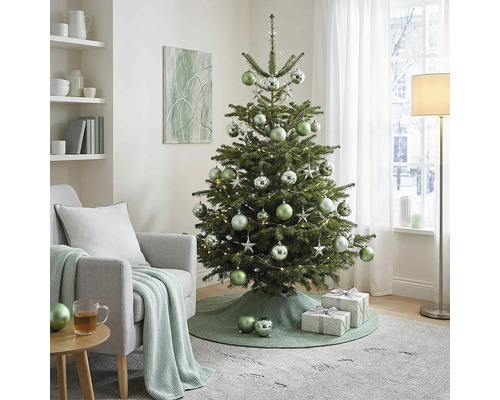 Geschmückter Weihnachtsbaum mit Kugeln und Lichtern im hellen Wohnzimmer neben einem Sessel mit Strickdecke und Geschenken.