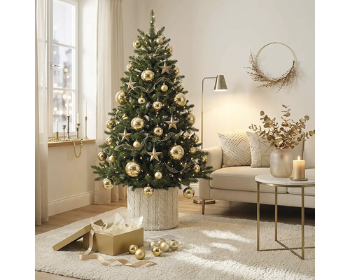 Geschmückter Weihnachtsbaum mit Sternen und Kugeln in einem hellen, organisierten Wohnzimmer. Der Baum steht in einer Strickhülle neben einem Sofa.
