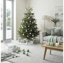 Festlich geschmückter Christbaum in einem hellen Wohnzimmer mit Sofa, Couchtisch aus Holz sowie silbernen und grünen Christbaumkugeln.