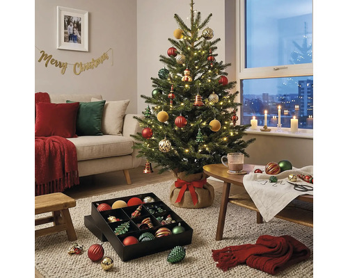 Dekorierter Christbaum mit Lichterkette in einem Wohnzimmer mit Sofa und einer Box voller Baumschmuck auf dem Teppich.