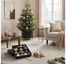 Festlich geschmückter Weihnachtsbaum im Metallständer in einem hellen Wohnzimmer mit Sofa, Couchtisch und Christbaumschmuck in einer Box.