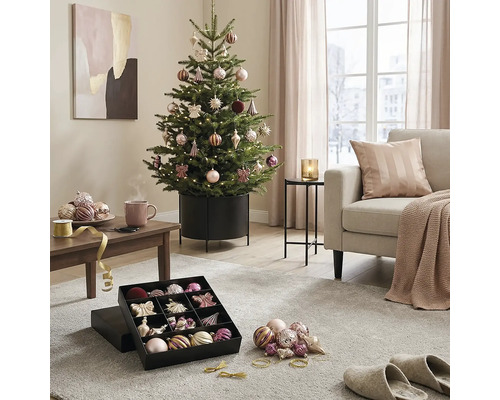 Festlich geschmückter Weihnachtsbaum im Metallständer in einem hellen Wohnzimmer mit Sofa, Couchtisch und Christbaumschmuck in einer Box.