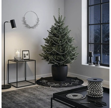 Modern eingerichtetes Zimmer mit einem Weihnachtsbaum im schwarzen Topf, dekoriert mit silbernen Perlenketten, Beistelltisch und Stehlampe.