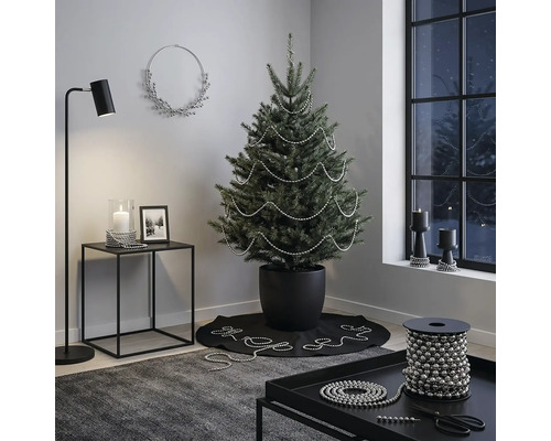 Modern eingerichtetes Zimmer mit einem Weihnachtsbaum im schwarzen Topf, dekoriert mit silbernen Perlenketten, Beistelltisch und Stehlampe.