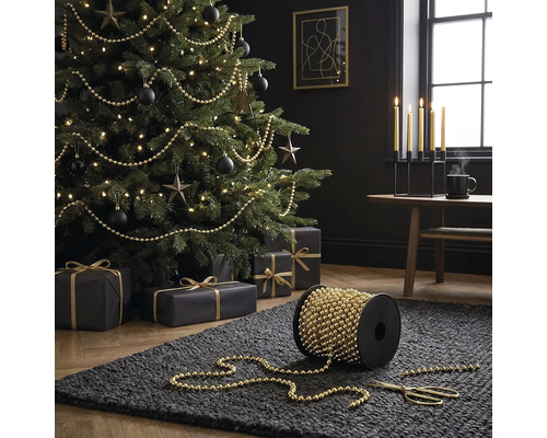 Weihnachtsbaum dekoriert mit goldfarbener Perlenkette, Spule mit Perlenkette im Vordergrund auf einem dunklen Teppich.