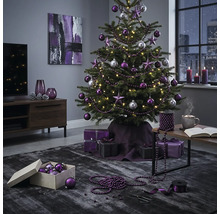 Festlich geschmückter Christbaum mit violetten und silbernen Kugeln sowie Sternen in einem modern eingerichteten Wohnzimmer mit Geschenken.