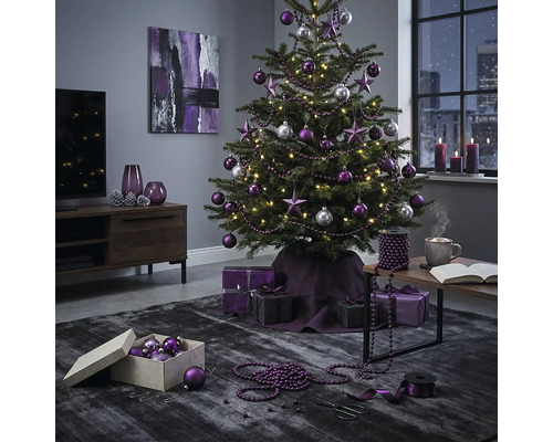 Festlich geschmückter Christbaum mit violetten und silbernen Kugeln sowie Sternen in einem modern eingerichteten Wohnzimmer mit Geschenken.