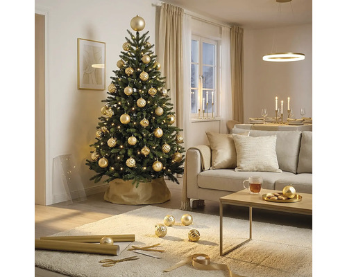 Festlich geschmückter künstlicher Christbaum mit goldenen Kugeln und Lichtern in einem hellen Wohnzimmer mit Sofa und Couchtisch.