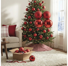 Geschmückter Christbaum mit roten matten und glänzenden Kugeln, warmweißen Lichtern und einer karierten Baumschürze im hellen Wohnzimmer.